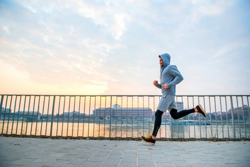 Person jogging along a waterfront path with metal railing and city buildings in background at sunrise or sunset