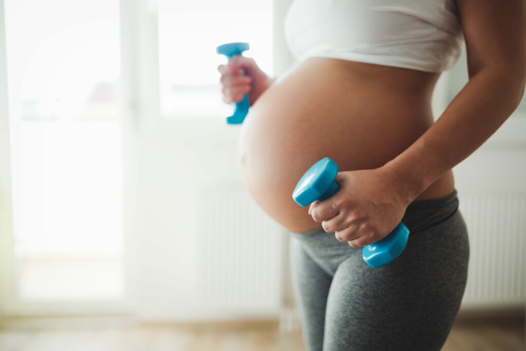 Pregnant woman holding blue dumbbells while exercising at home near a bright window