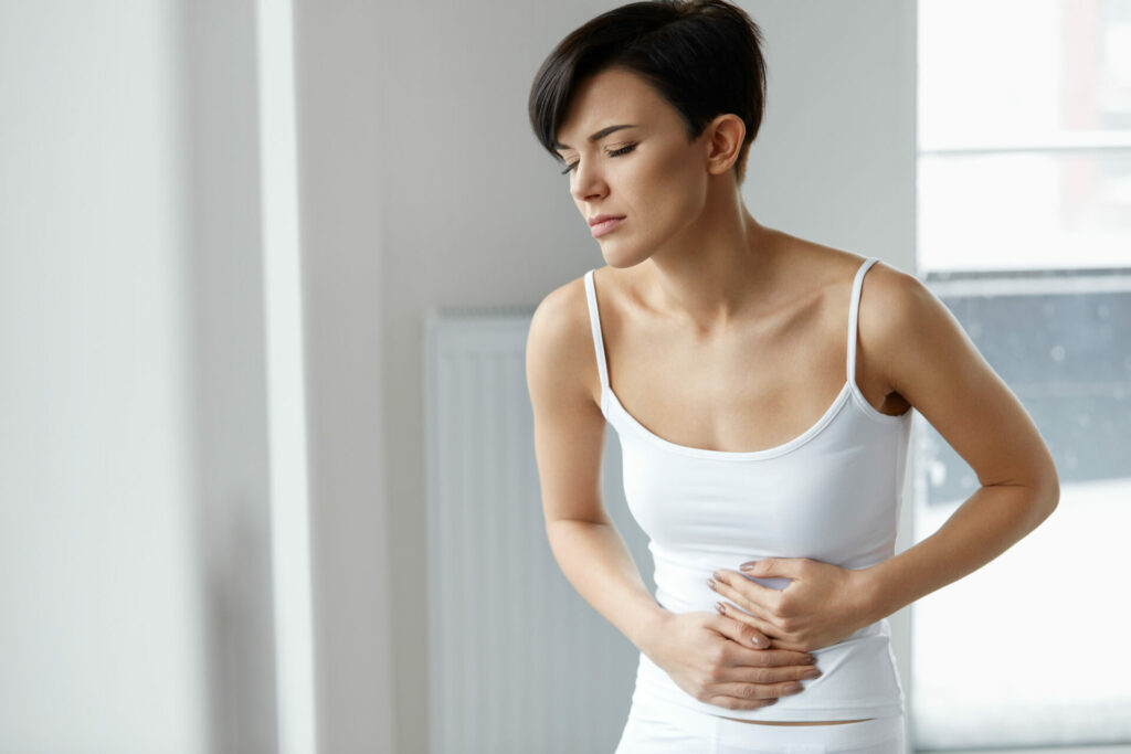 Woman in white tank top holding her stomach in discomfort while looking down