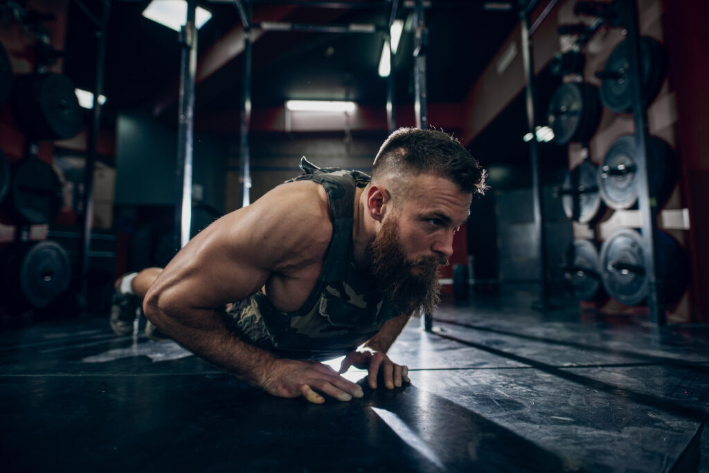 Bearded man performing a plank exercise on dark gym floor with equipment visible in background
