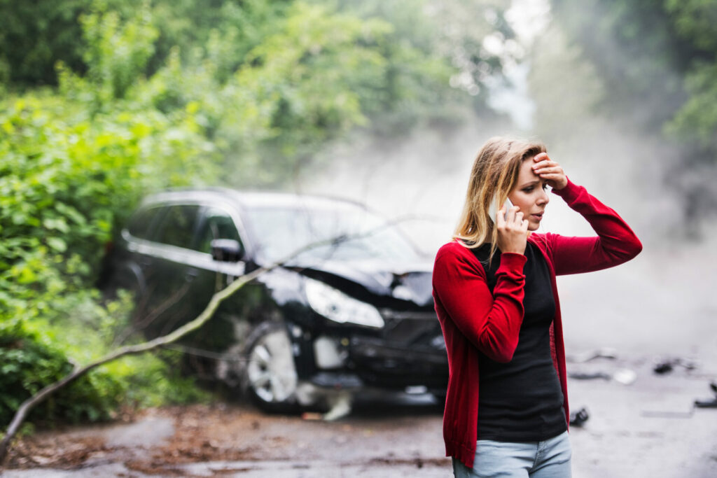 Woman in red cardigan on phone with damaged black car and smoke in background