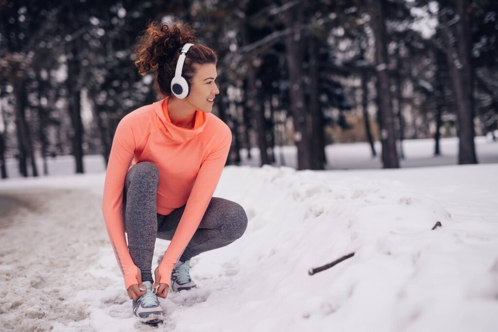 Woman in orange long-sleeve shirt and gray leggings stretching outdoors in snowy forest while wearing white headphones