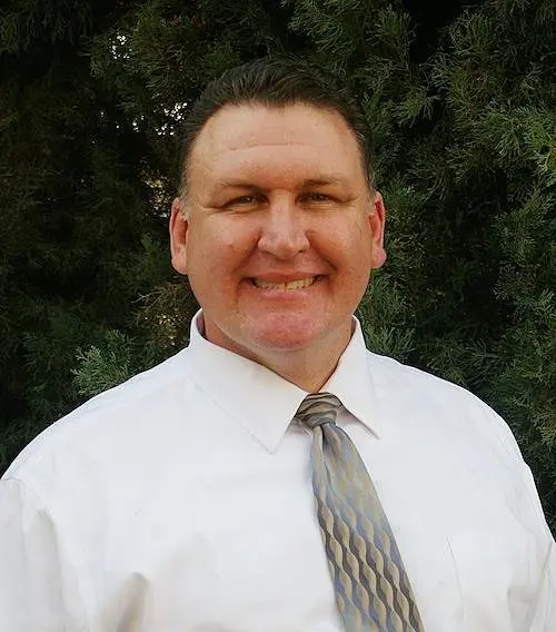 Dr. Raymond Espinosa in a white shirt and patterned tie, smiling, with evergreen trees behind him
